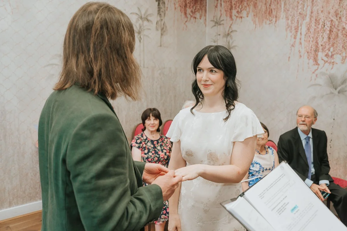  Bride and groom exchanging wedding rings in the  Pauline Room at Lambeth Town Hall , surrounded by close family and friends during their London registry office ceremony. 