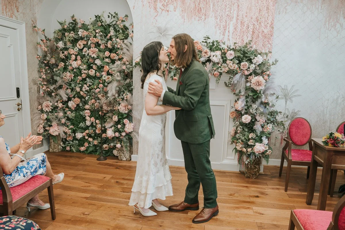  Newly married couple sharing their first kiss in the  Pauline Room at Lambeth Town Hall , celebrating their marriage beneath the room's beautiful floral display. 