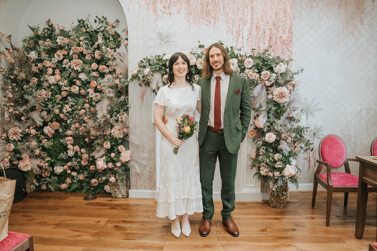  Bride and groom posing together in the  Pauline Room at Lambeth Town Hall , with the elegant floral ceremony backdrop after their intimate London registry office wedding. 