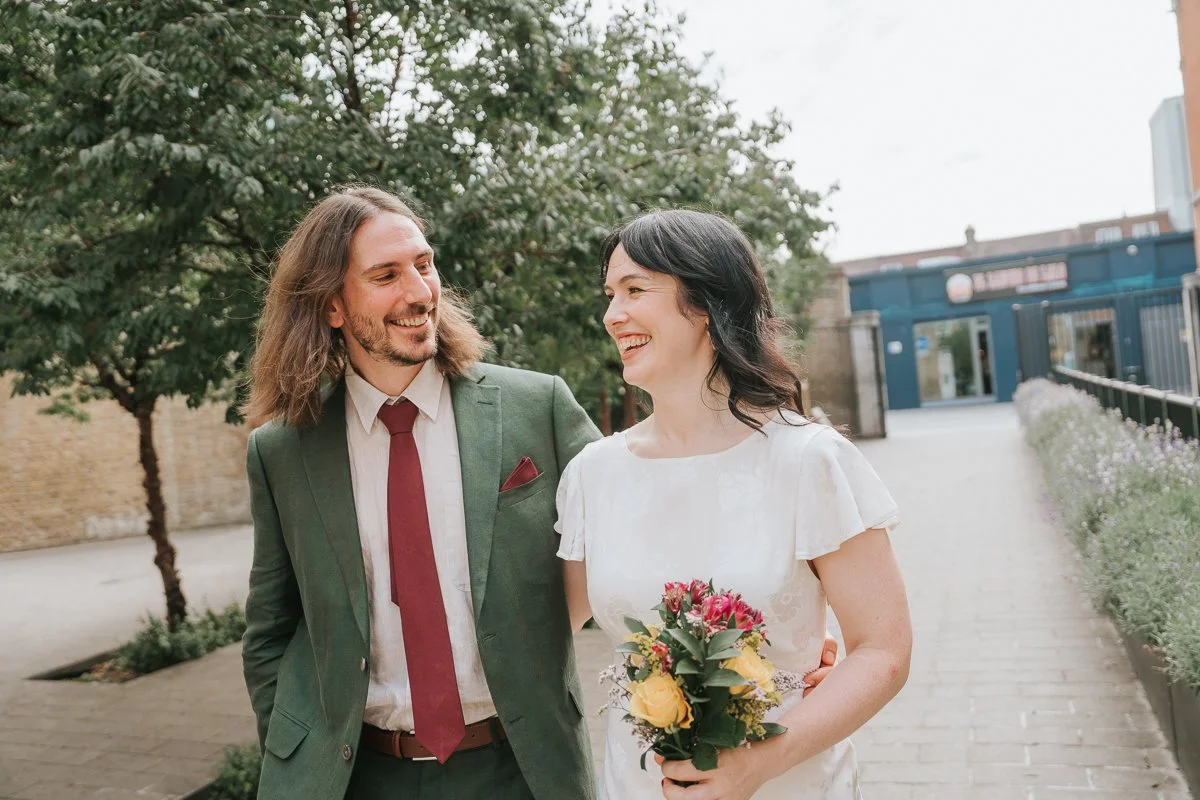  Bride and groom walking arm in arm outside  Lambeth Town Hall in Brixton , smiling at each other as they stroll through the landscaped courtyard after their intimate London registry office wedding, with the bride holding a colourful bouquet. 
