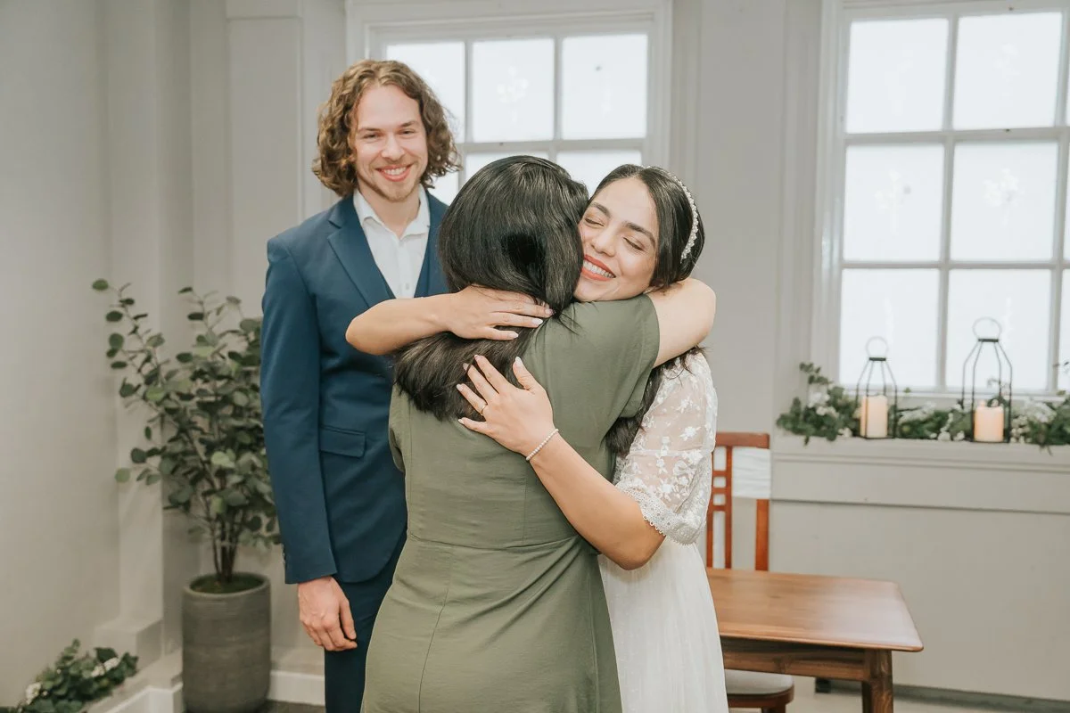  Bride embracing a family member after the ceremony while the groom smiles during their Kingston Register Office wedding. 