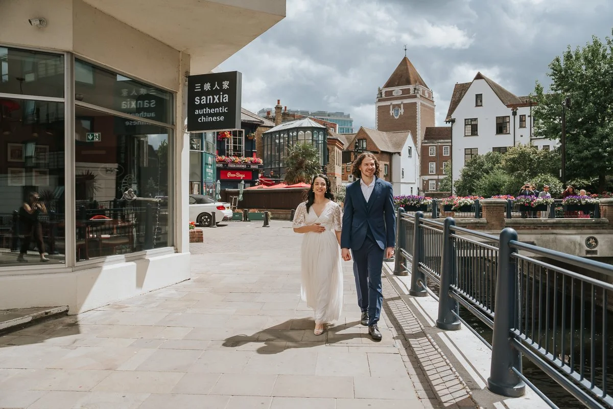  Bride and groom walking hand in hand across Kingston riverside footbridge after their Kingston Register Office wedding, laughing together during relaxed couple portraits. 
