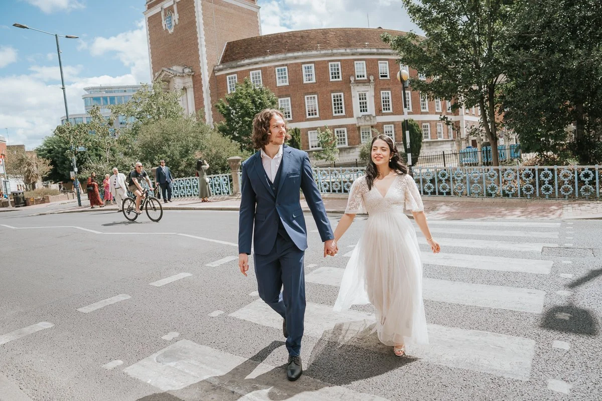  Bride and groom walking hand in hand across Kingston riverside footbridge after their Kingston Register Office wedding, laughing together during relaxed couple portraits. 