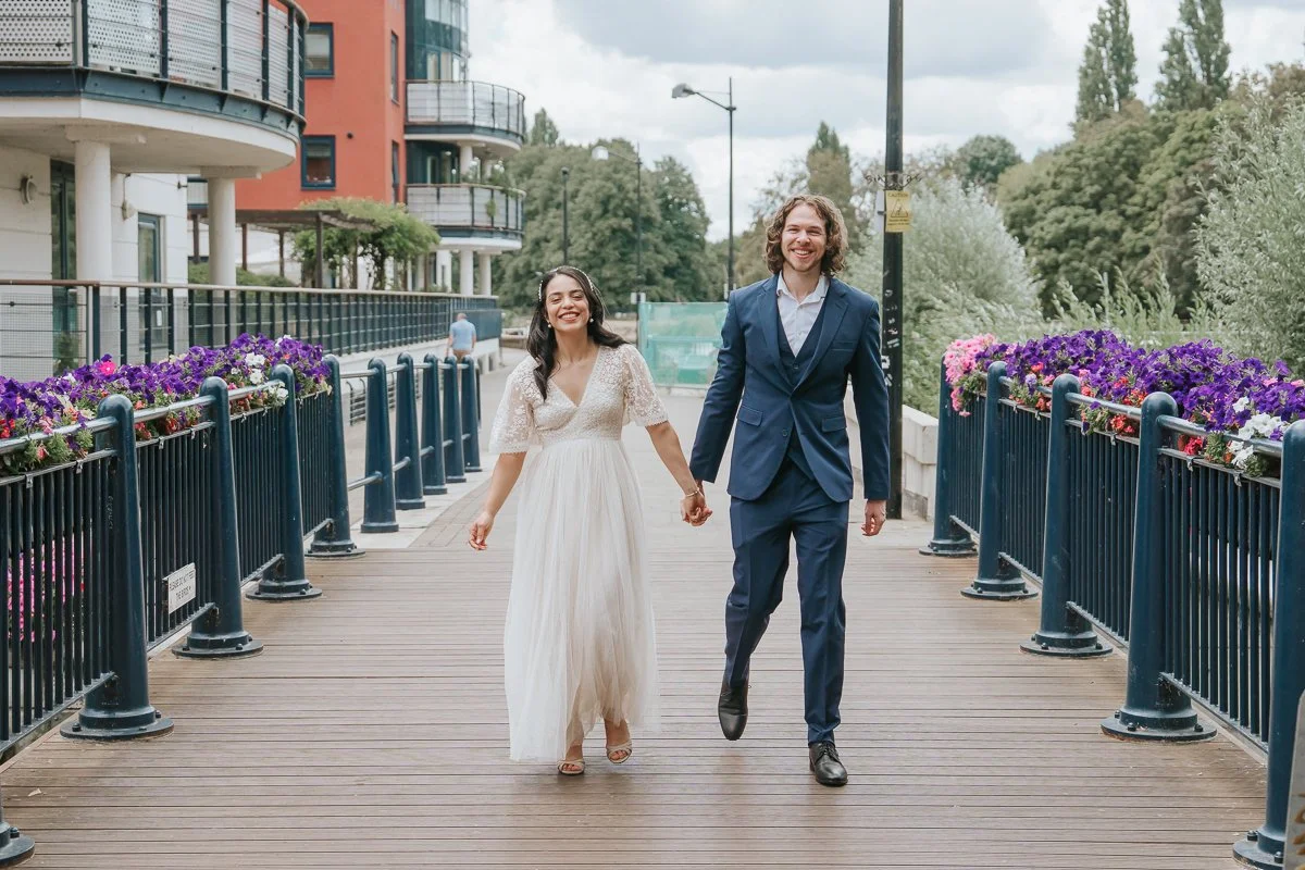  Newly married couple strolling along Kingston riverside surrounded by colourful flowers, capturing natural wedding photography in Kingston upon Thames. 