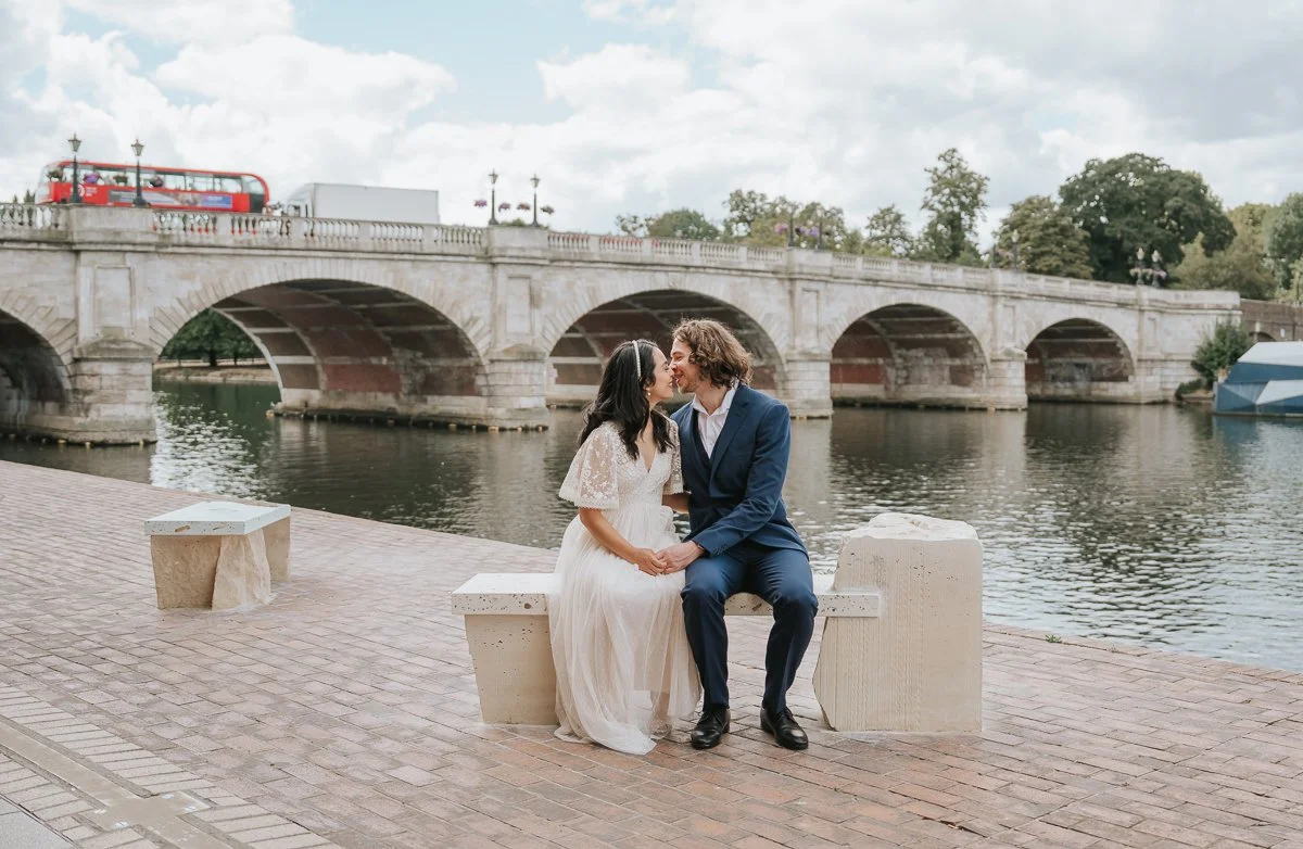  Bride and groom sitting together by the River Thames beneath Kingston Bridge during a relaxed wedding photoshoot in Kingston upon Thames. 