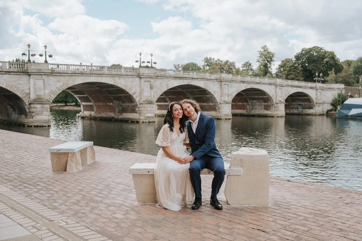 Bride and groom sitting together by the River Thames beneath Kingston Bridge during a relaxed wedding photoshoot in Kingston upon Thames. 