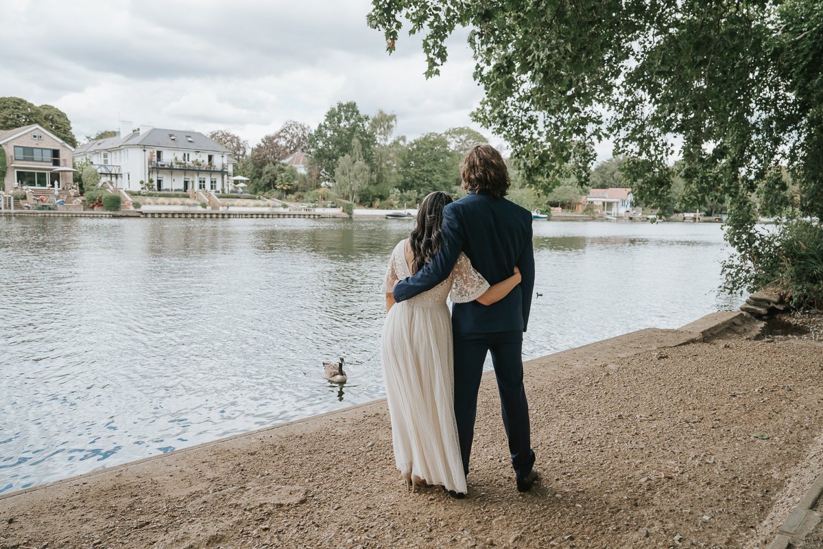  Romantic back view of the bride and groom embracing beside the River Thames during their Kingston riverside wedding photoshoot. 
