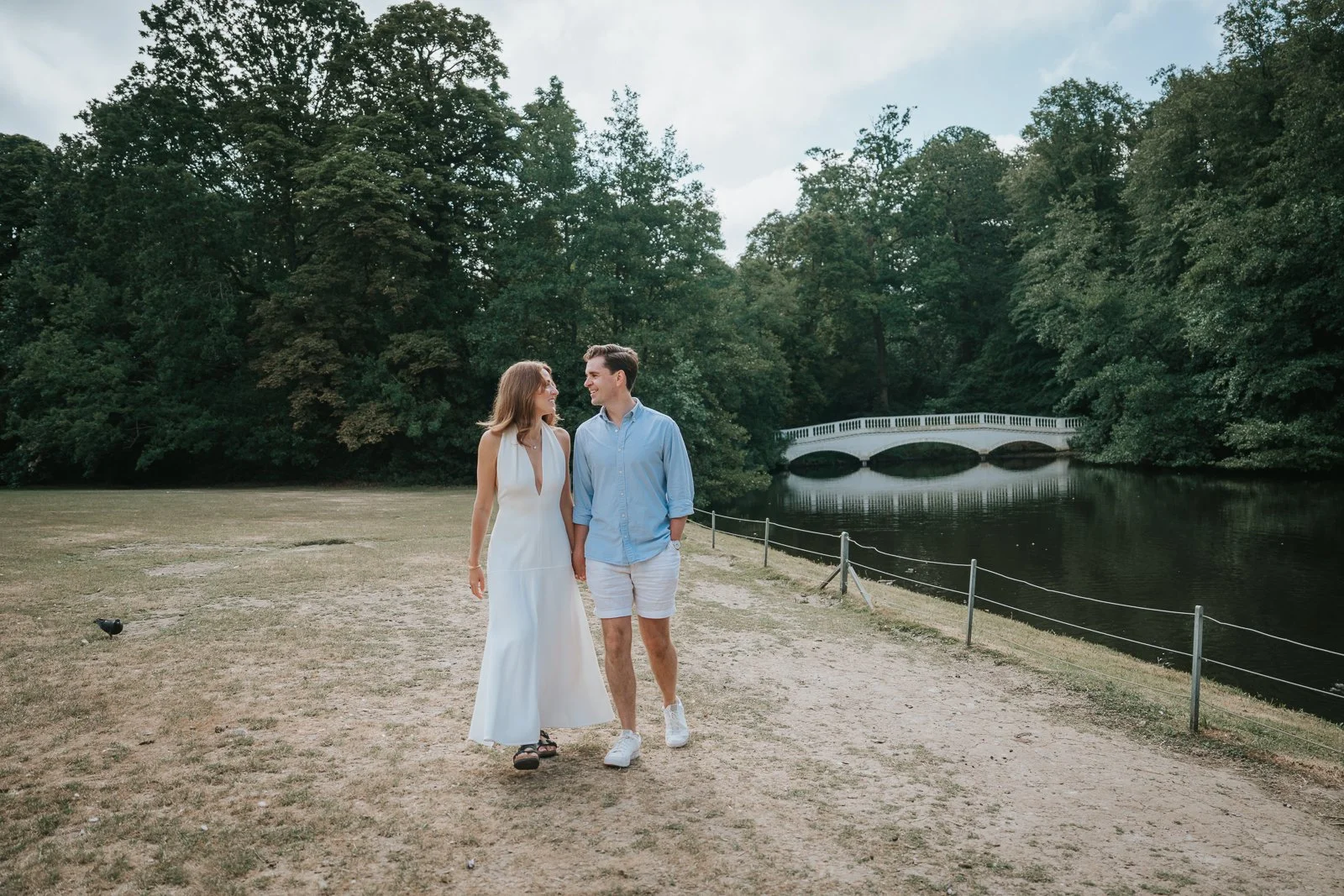  Couple walking hand in hand beside the lake at Hampstead Heath with the iconic white bridge near Kenwood House in the background during a relaxed engagement photoshoot. 