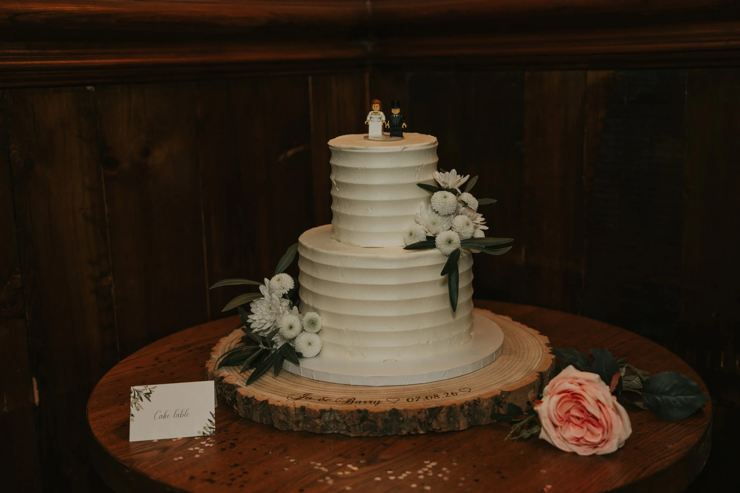  Two-tier white wedding cake decorated with flowers and a LEGO bride and groom topper at the County Arms in Wandsworth. 