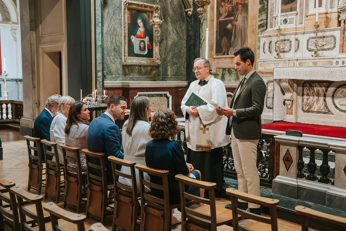  Intimate family christening ceremony at Brompton Oratory, with the priest and godparent beside the ornate altar. 