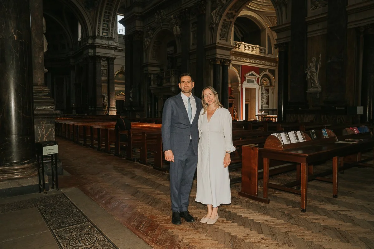  Parents standing inside Brompton Oratory before their baby’s christening, surrounded by the church’s grand historic interior. 