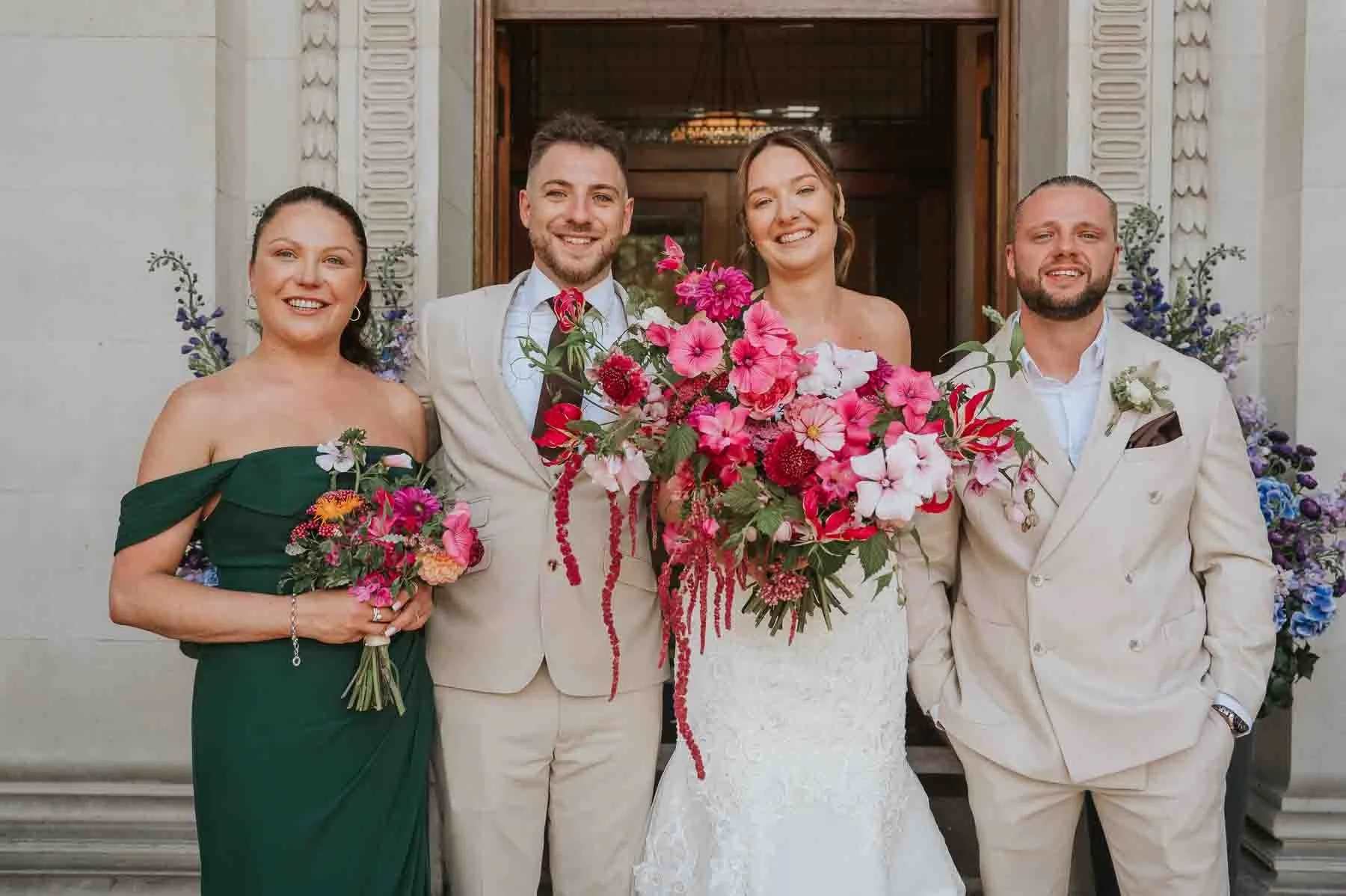  Marylebone Town Hall wedding photography of the newlyweds with their wedding party outside the entrance, surrounded by colourful floral arrangements. 