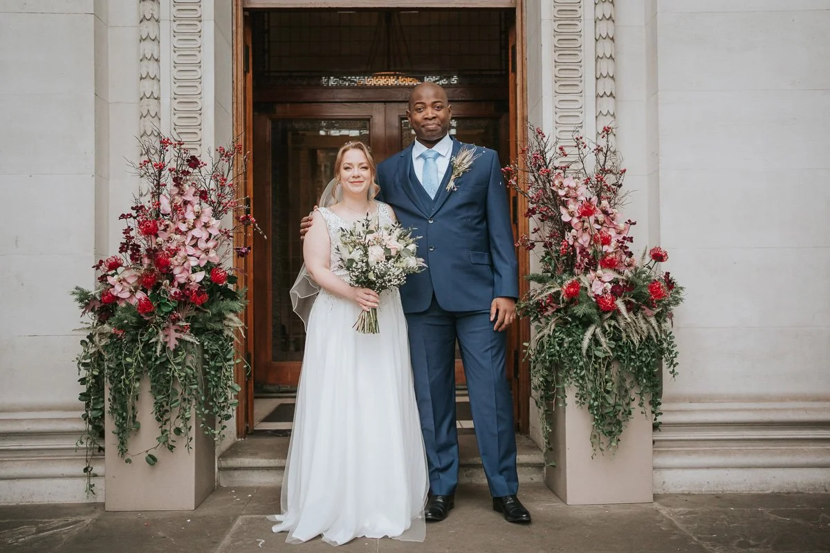   Bride and groom posing outside Marylebone Town Hall after their wedding ceremony, framed by beautiful floral arrangements at the entrance.  