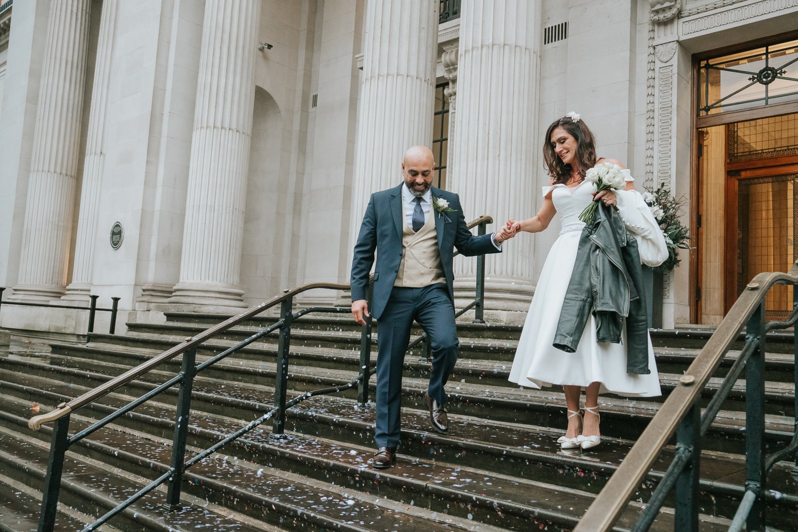  Newly married couple walking down the steps of Old Marylebone Town Hall, holding hands, iconic London registry office wedding photo. 