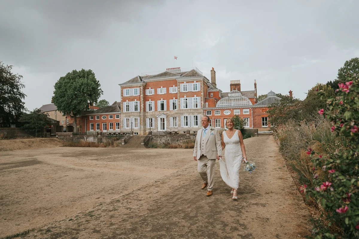  Bride and groom walking through the grounds of York House in Twickenham, with the historic wedding venue and gardens creating a striking backdrop. 