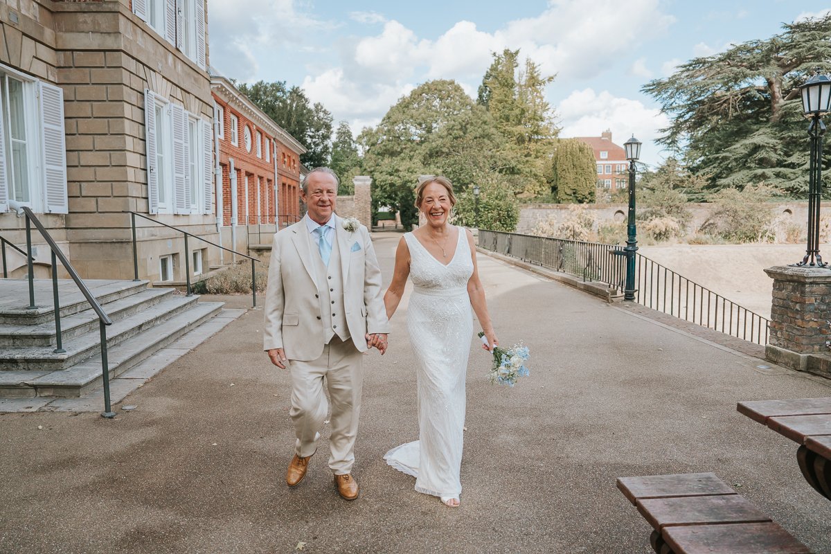  Bride and groom walking hand in hand beside York House in Twickenham, captured during their relaxed wedding photography in the grounds. 