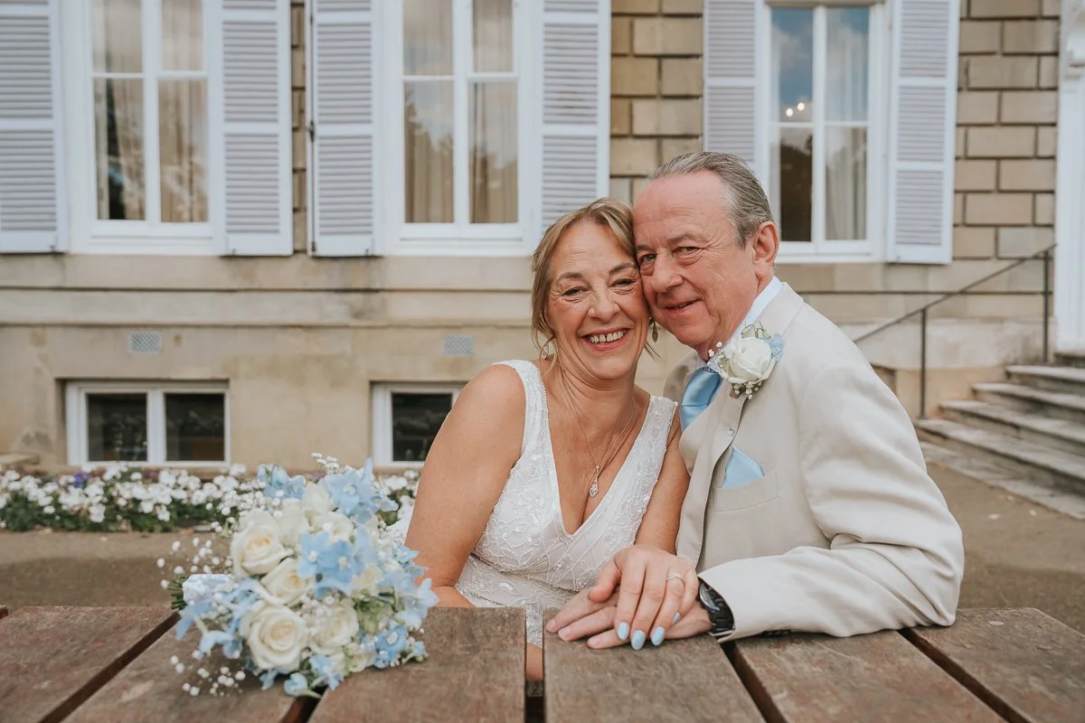  Bride and groom smiling together outside York House in Twickenham, with a blue and white wedding bouquet in the foreground. 