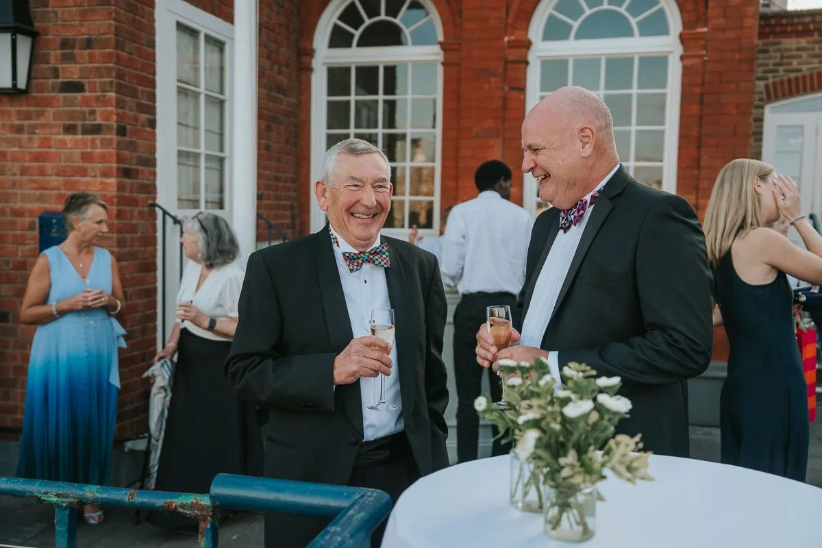  Two guests laughing over champagne at an outdoor birthday party at The Queen’s Club. 