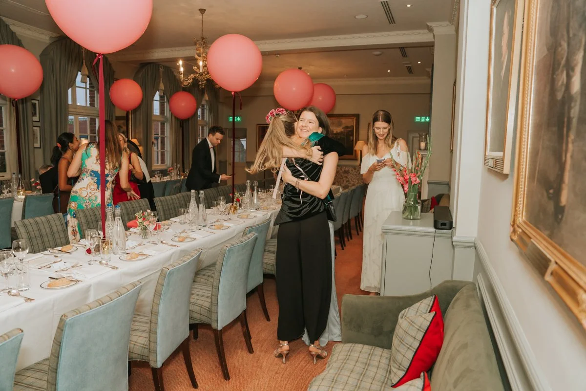  Friends greeting with a hug in the pink balloon-decorated dining room at The Queen’s Club. 