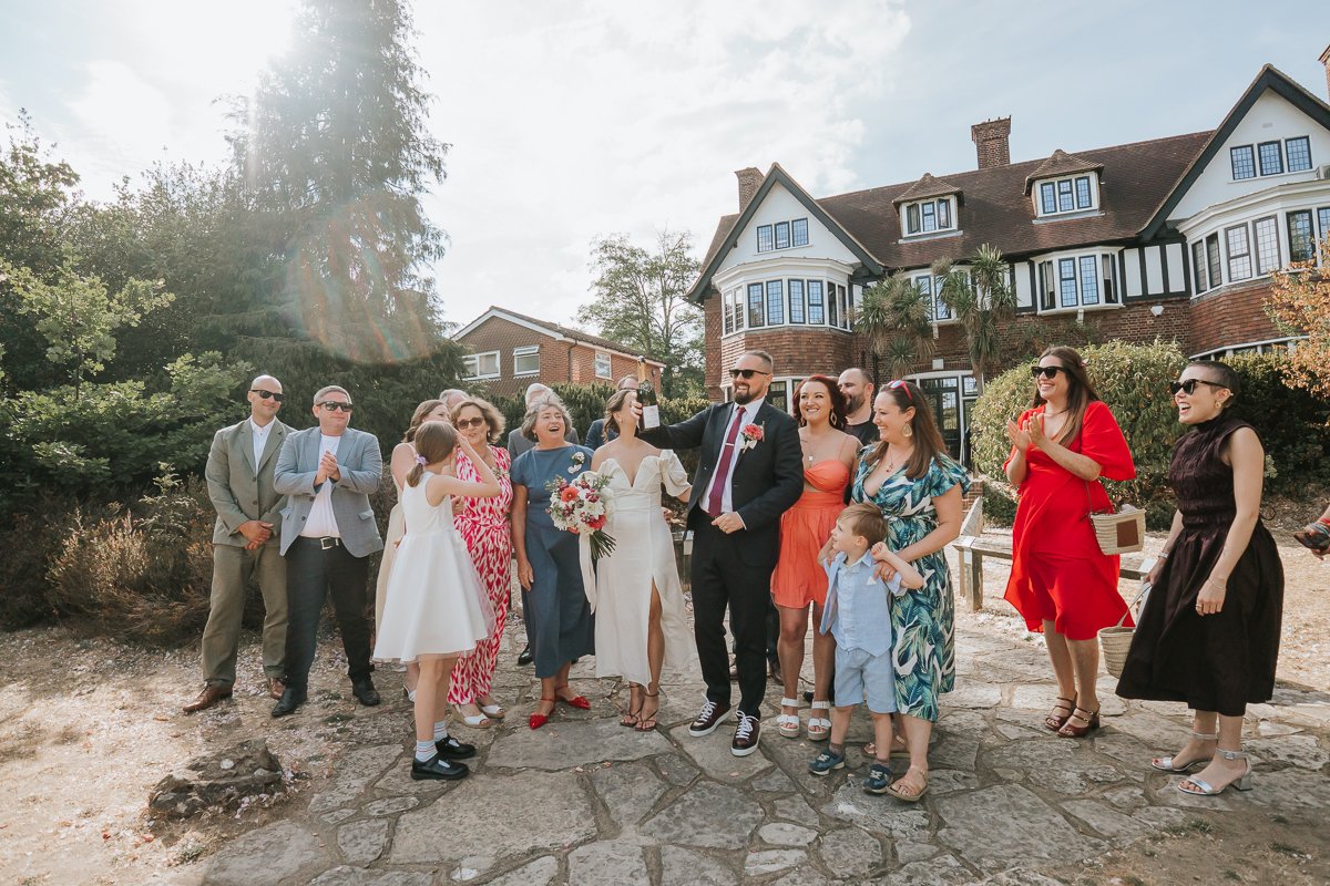  Relaxed group wedding photo of the bride and groom with their guests in the grounds of Weybridge Register Office. 