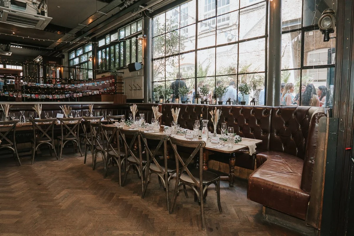  The Depot Islington wedding reception set-up with long dining tables beside the venue’s large windows and leather seating. 
