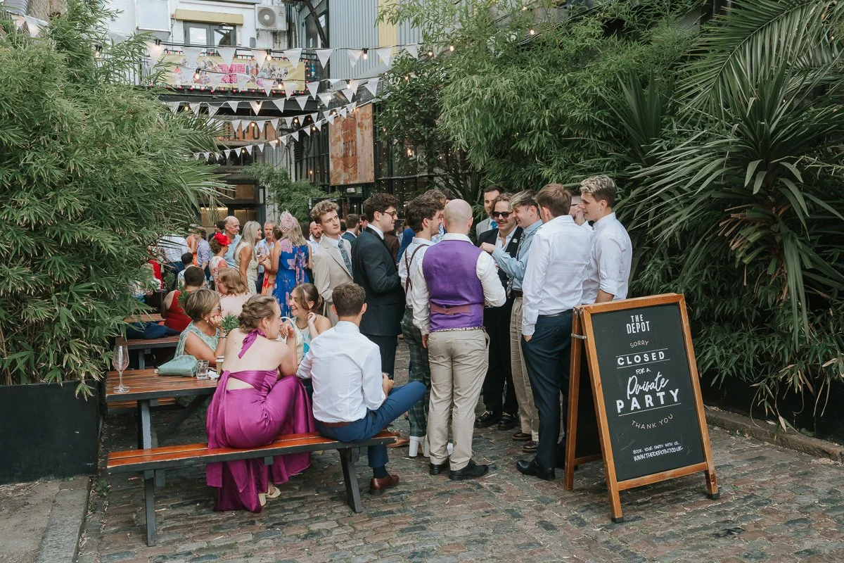 Wedding guests enjoying drinks and chatting in the leafy courtyard at The Depot in Islington, London. 
