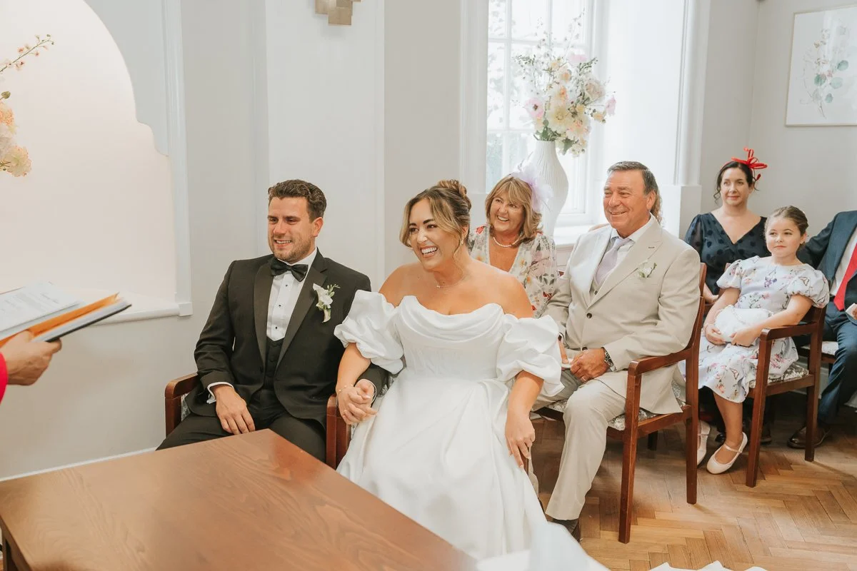  Bride and Groom sit next to each other holding hands as they listen to the registrar speak during their wedding at The Rossetti Room in Chelsea Old Town Hall. 