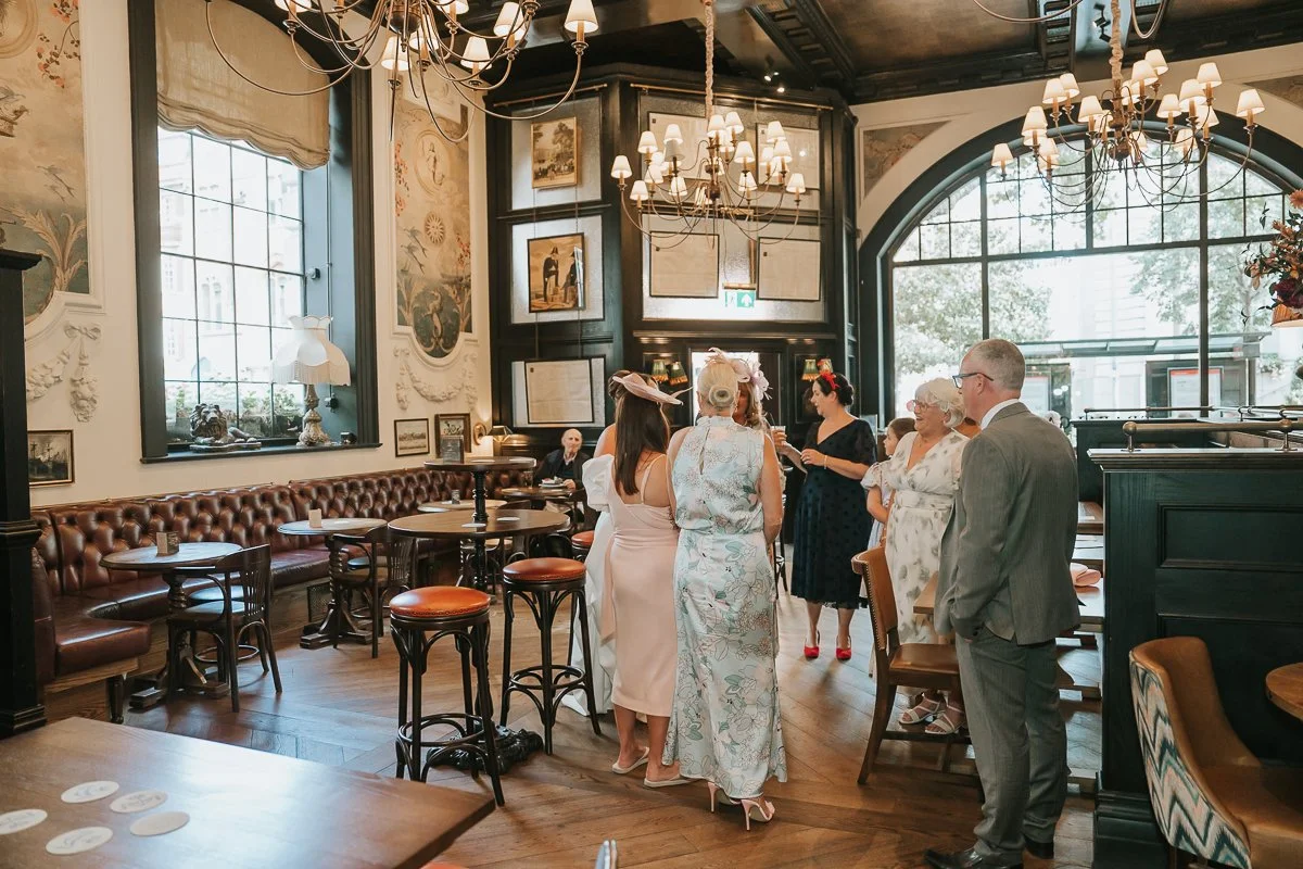  Wedding guests chatting beside the large arched windows and leather seating inside The Trafalgar Chelsea. 