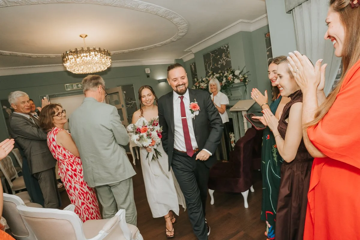  Bride and groom leaving their Weybridge Register Office wedding ceremony together, surrounded by smiling guests. 