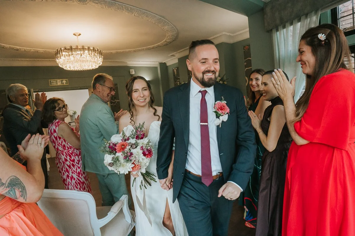  Bride and groom leaving their Weybridge Register Office wedding ceremony together, surrounded by smiling guests. 