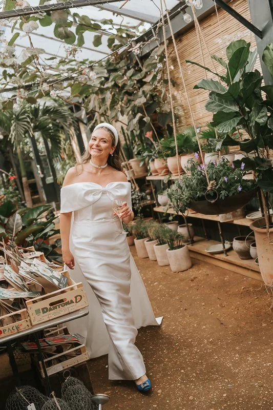  Bride holding a drink and smiling among potted plants and hanging baskets inside the greenhouse at Petersham Nurseries, Richmond. 
