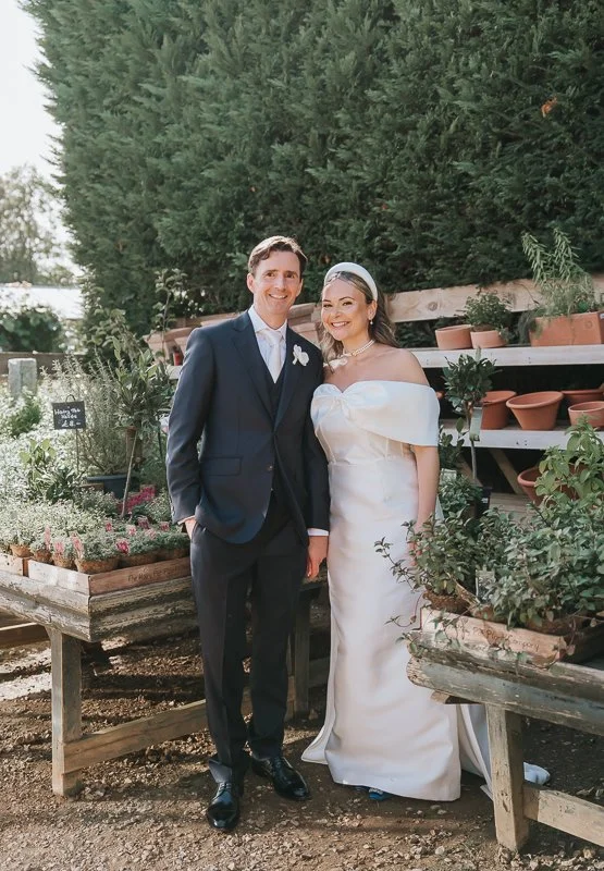  Bride and groom smiling among outdoor plant displays and terracotta pots at Petersham Nurseries in Richmond. 