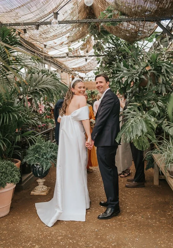  Bride and groom holding hands and looking back at the camera inside the leafy greenhouse at Petersham Nurseries, Richmond. 