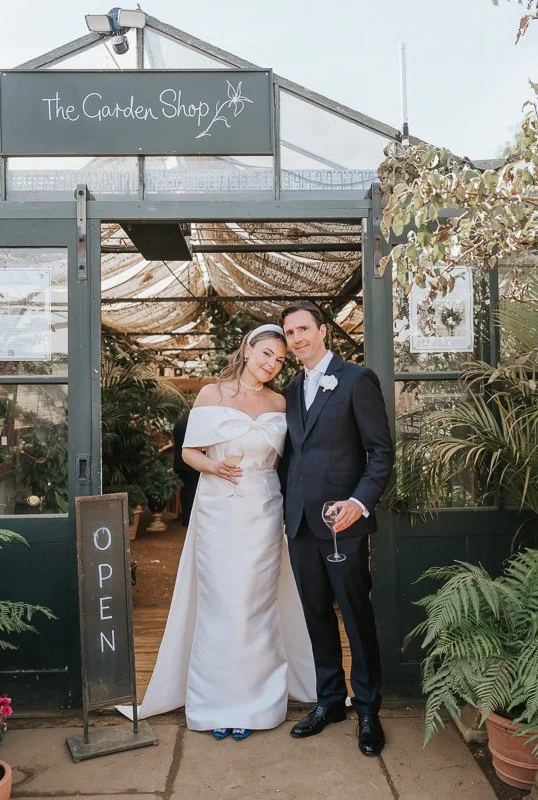  Bride and groom holding drinks outside the Garden Shop greenhouse, surrounded by plants at Petersham Nurseries in Richmond. 