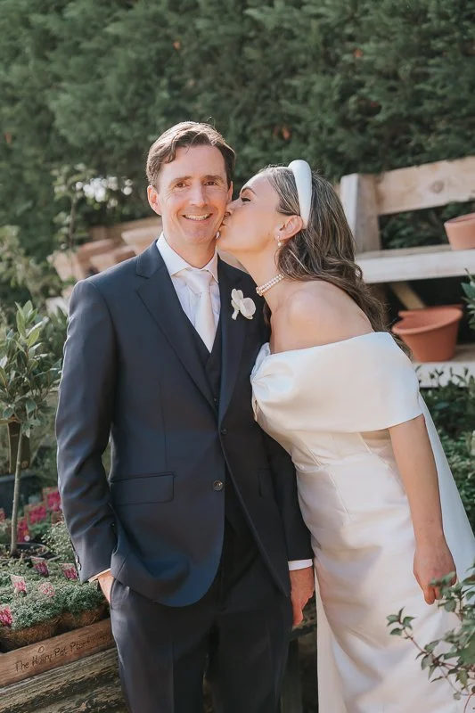  Bride kissing the groom’s cheek beside outdoor plant displays at Petersham Nurseries, Richmond. 