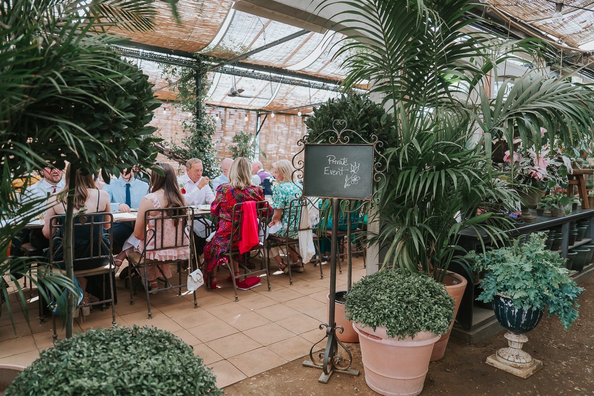  Wedding guests chatting around a table among greenhouse plants beside a private event sign at Petersham Nurseries, Richmond. 