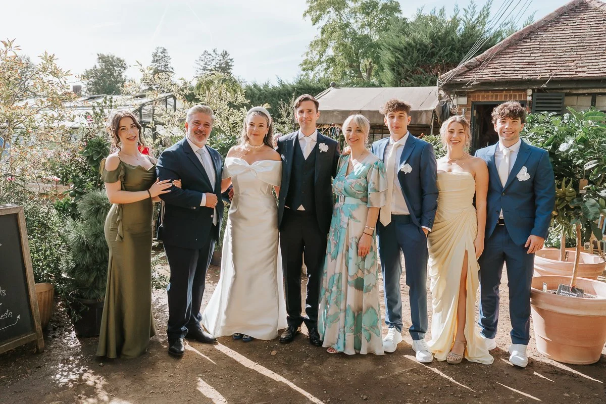  Bride and groom posing with six wedding guests among outdoor plants and terracotta pots at Petersham Nurseries, Richmond. 