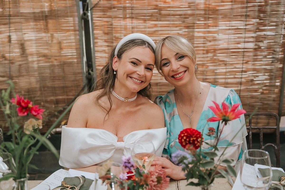  Bride and a wedding guest smiling together at a flower-filled table in the Central Greenhouse at Petersham Nurseries. 