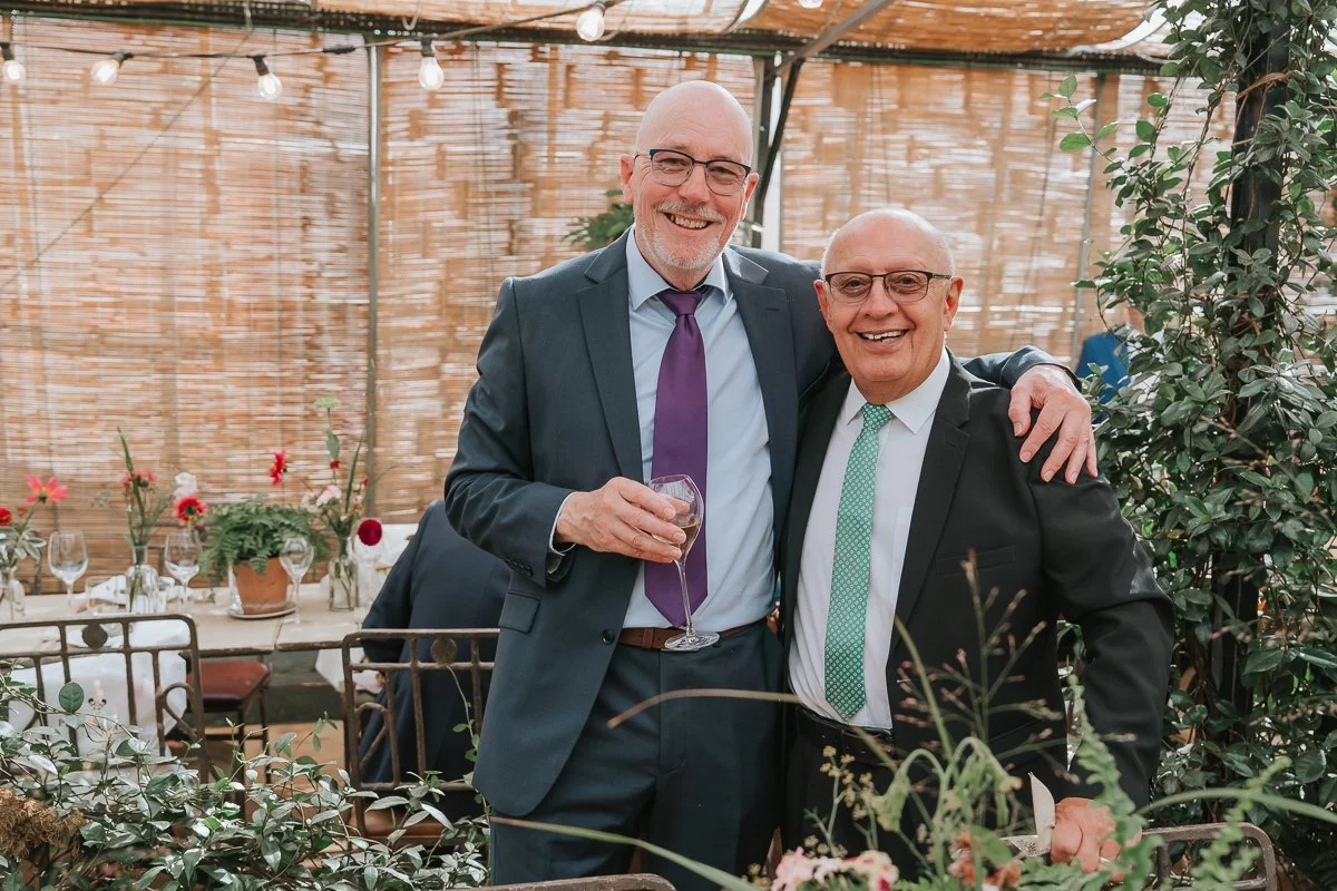  Two wedding guests smiling with their arms around each other beneath festoon lights in the Central Greenhouse at Petersham Nurseries. 