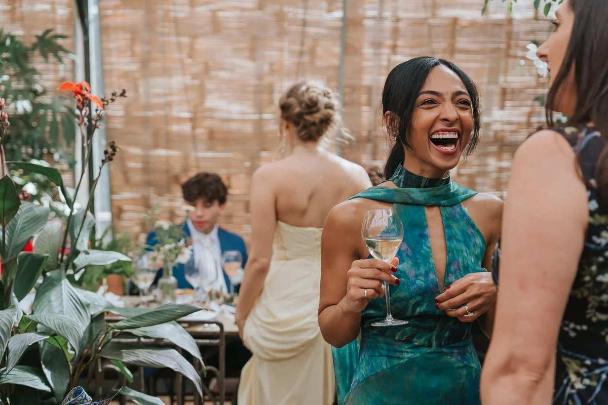  Wedding guest holding a glass of wine and laughing during the reception in the Central Greenhouse at Petersham Nurseries. 