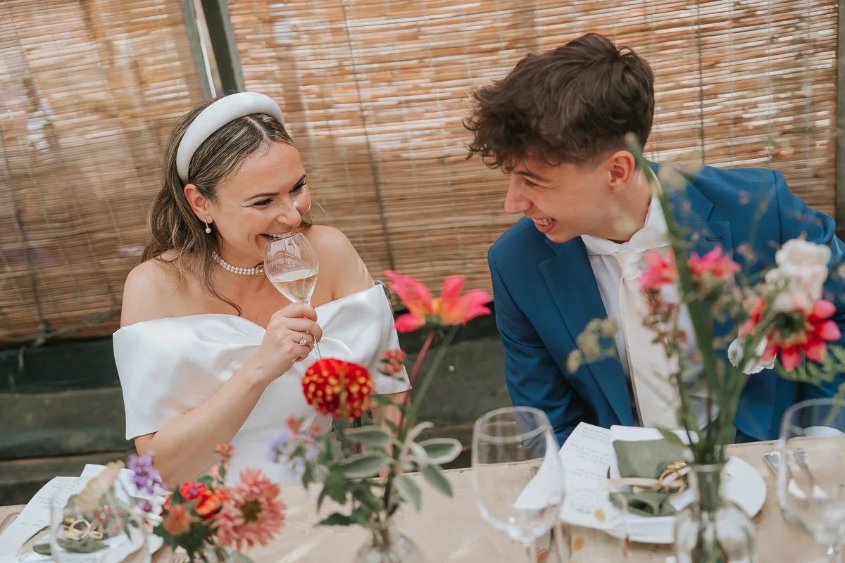  Bride laughing with a guest in a blue suit at their flower-filled wedding breakfast table at Petersham Nurseries. 