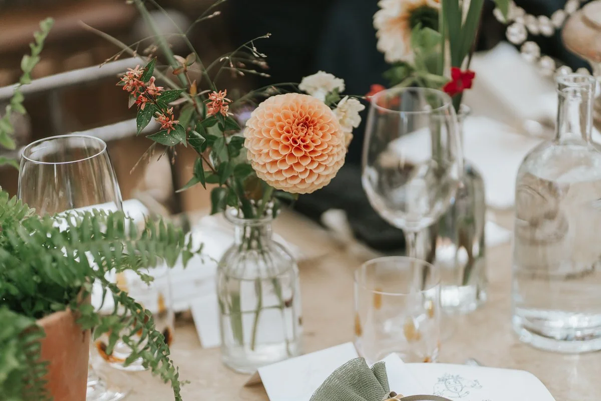  Peach dahlia in a glass bud vase beside wine glasses and fern foliage on a wedding table at Petersham Nurseries. 