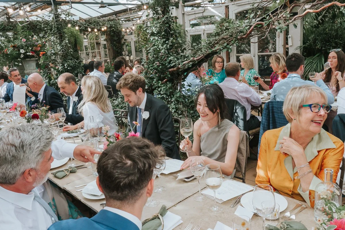  Wedding guests chatting at long, flower-filled tables beneath climbing greenery in the Central Greenhouse at Petersham Nurseries. 