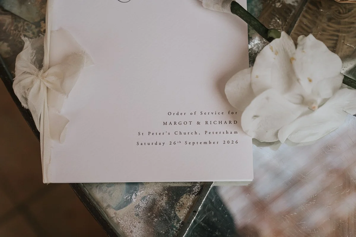  Close-up of Margot and Richard’s wedding order of service for St Peter’s Church, Petersham, beside white orchids. 