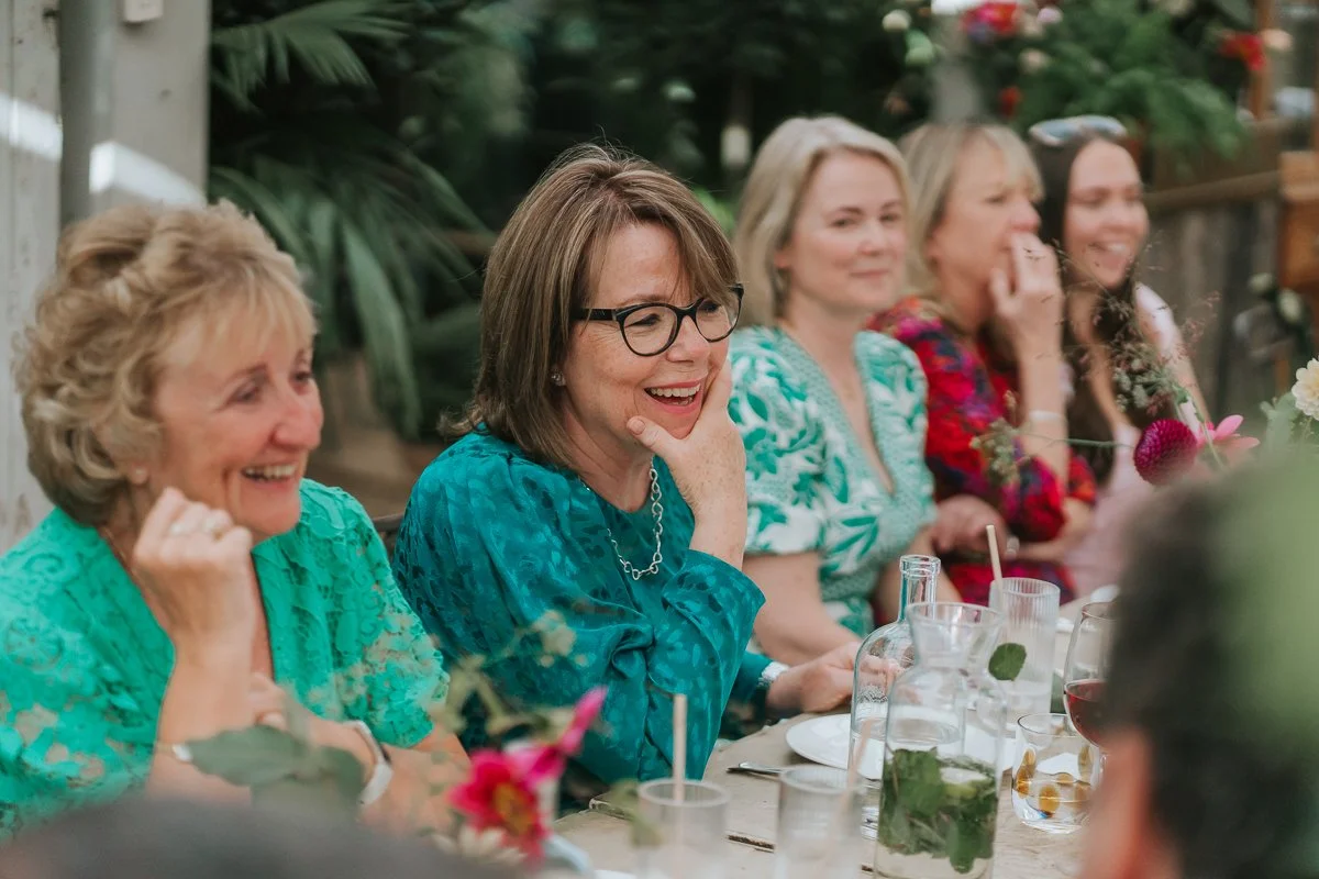  Wedding guests laughing during the speeches in the Central Greenhouse at Petersham Nurseries. 