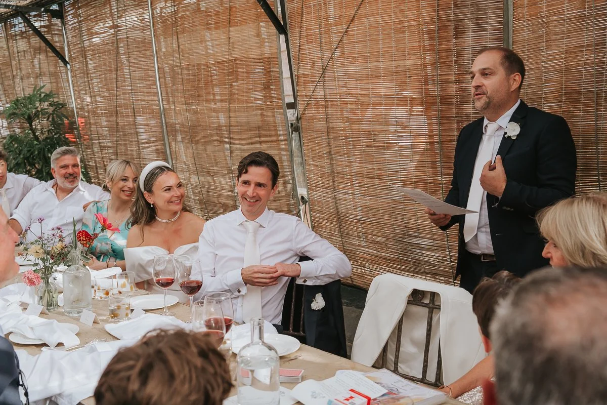  Guest giving a wedding speech as the bride and groom smile at their table in the Central Greenhouse. 