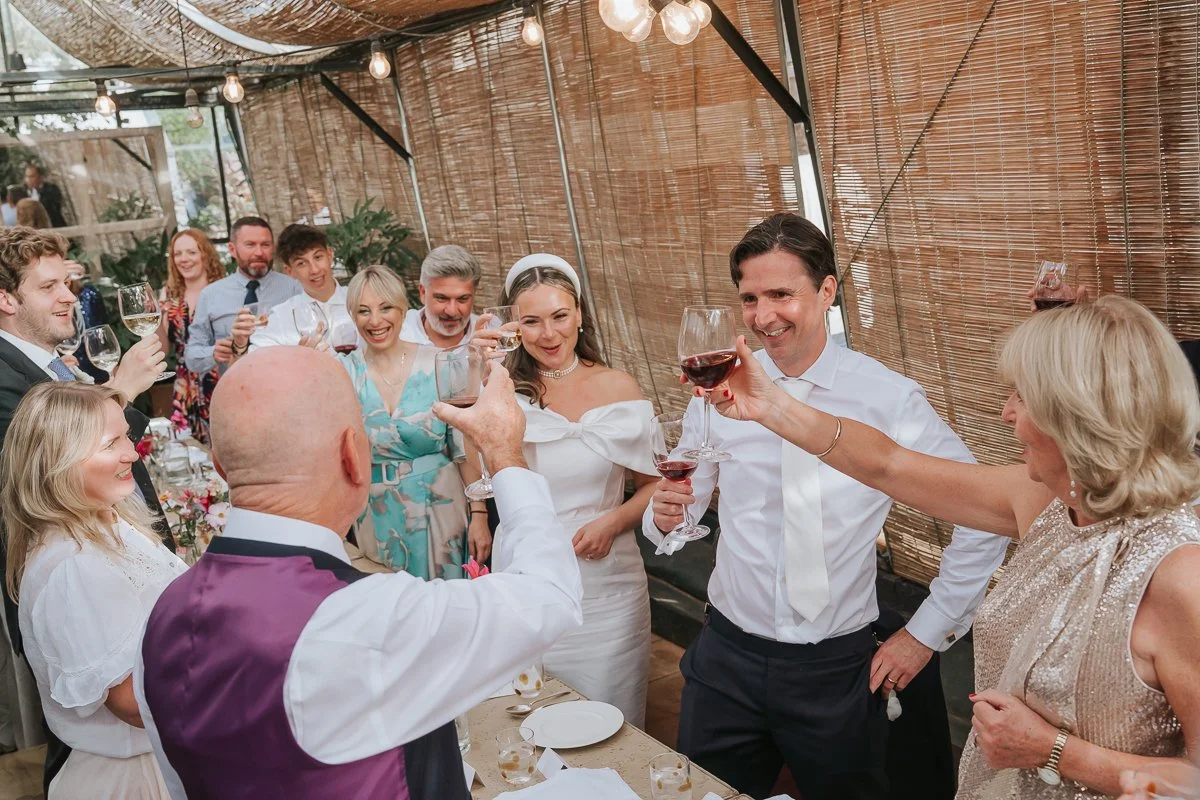  Bride, groom and guests raising their wine glasses for a wedding toast at Petersham Nurseries, Richmond. 