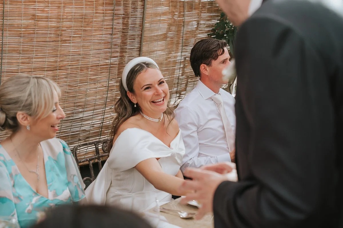  Bride smiling at a standing guest while seated beside the groom during their Petersham Nurseries wedding reception. 