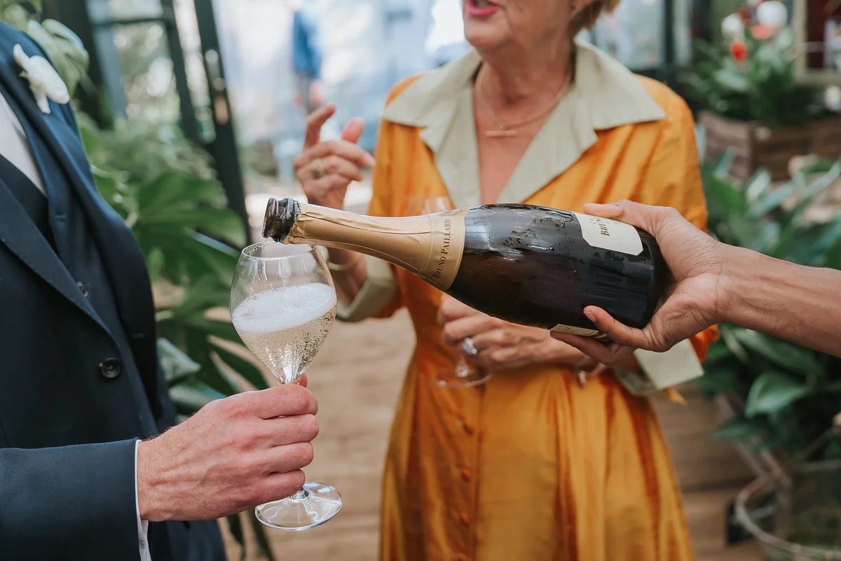  Champagne being poured into the groom’s glass during a wedding reception in the Central Greenhouse at Petersham Nurseries. 