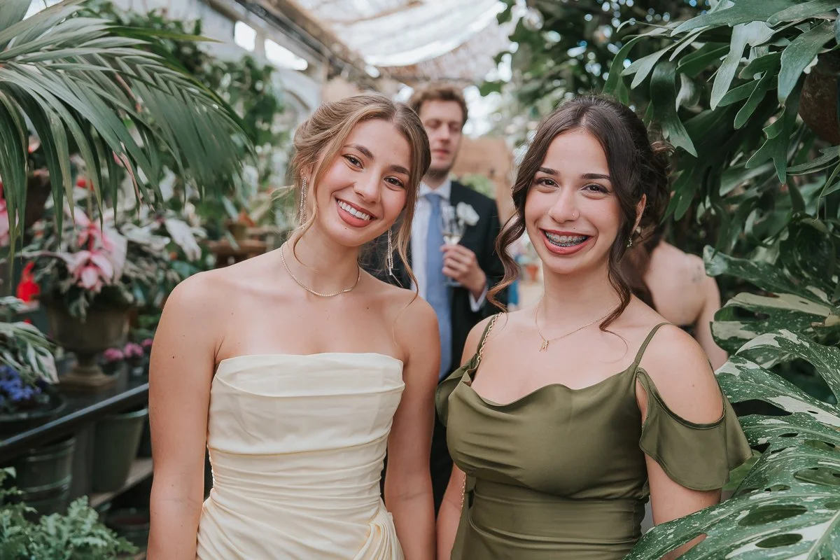  Two wedding guests in cream and olive-green dresses smiling among greenhouse plants at Petersham Nurseries 